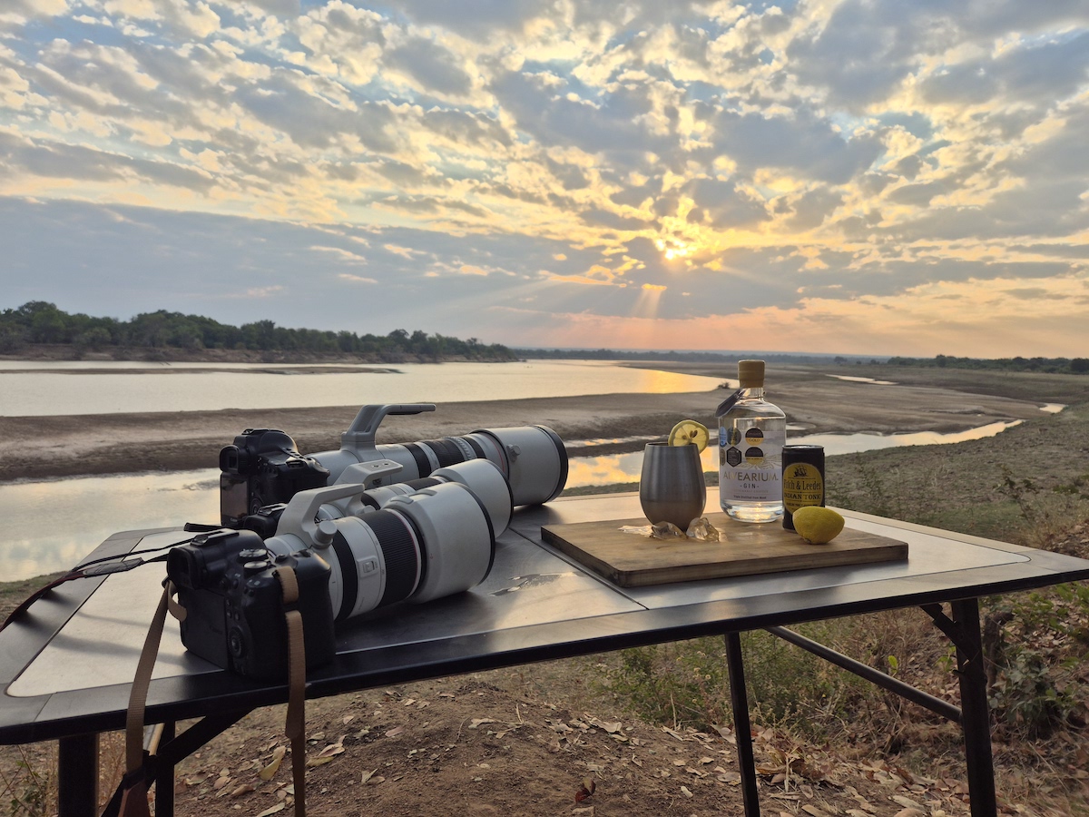 A camp table with cameras, gin and a river view at golden hour