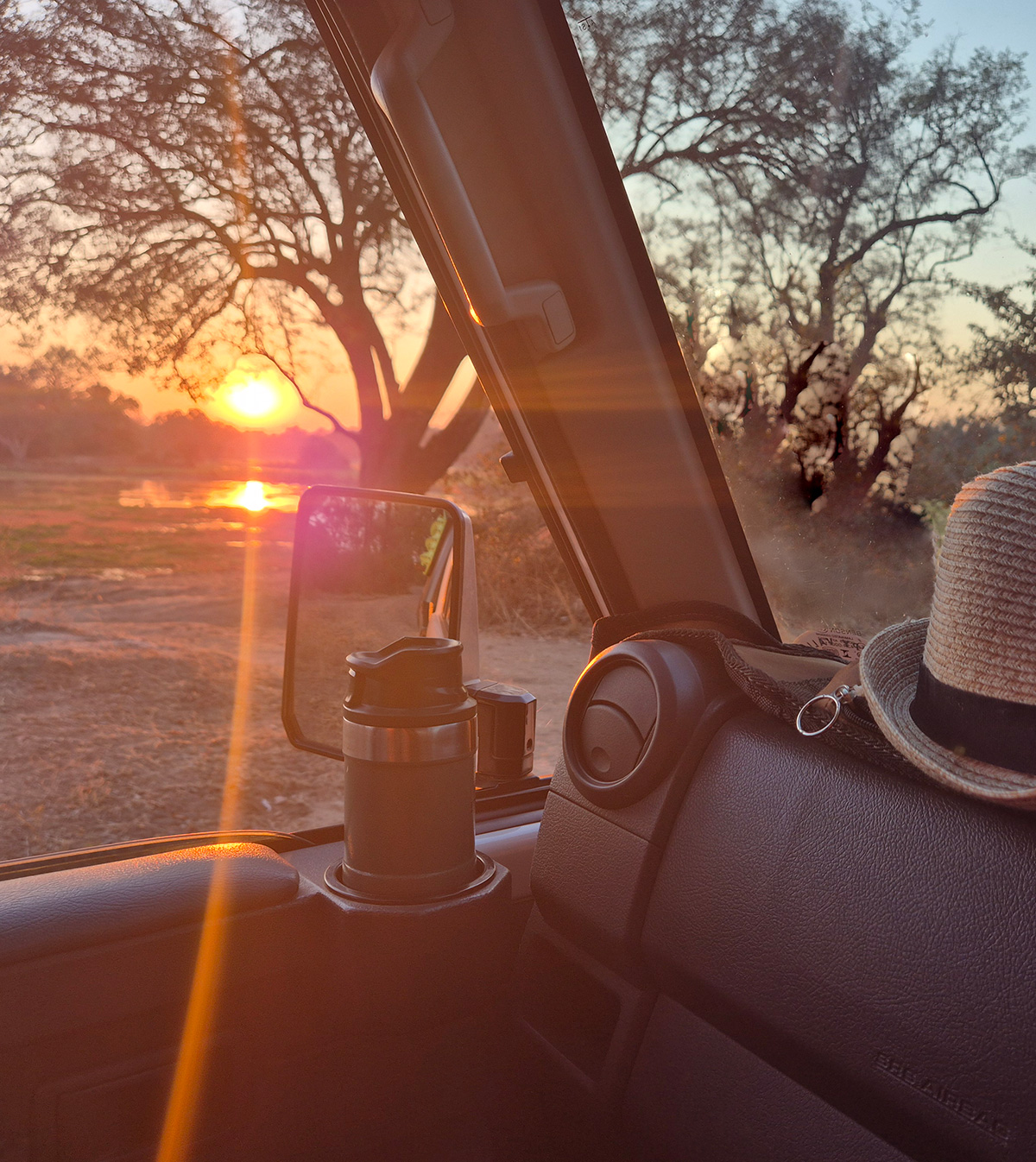 Sunset over the Zambian bush from the passenger seat of a 4x4, with a straw hat and travel mug on the dash